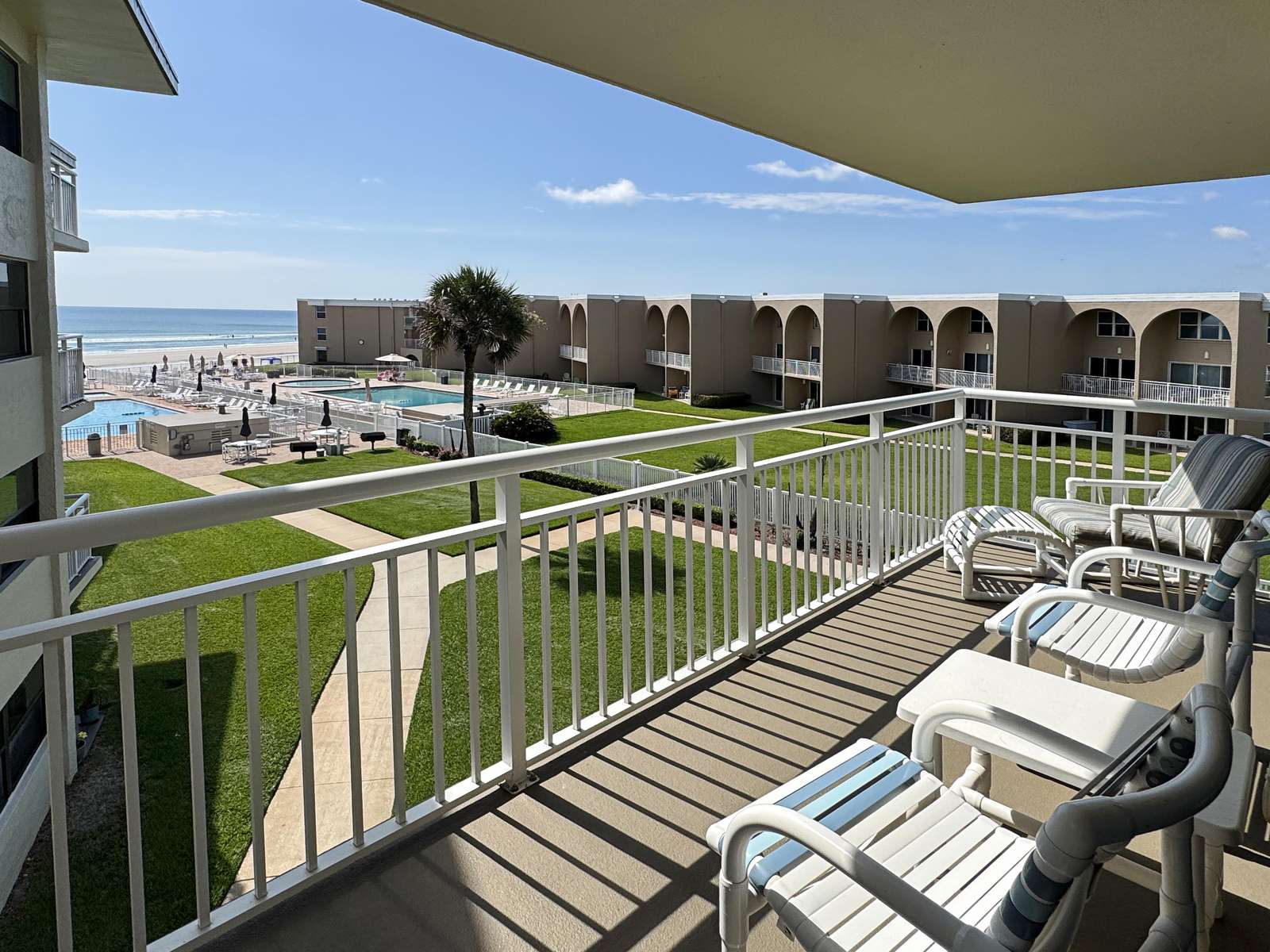 Balcony with white metal railing, striped cushioned chairs, and views of pool, lawn, and ocean beyond. - property