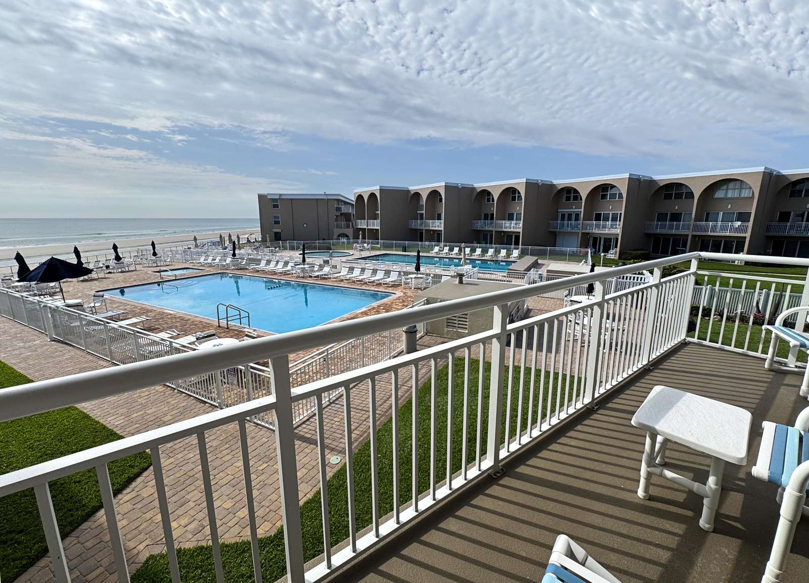 Balcony with white railing overlooks the pool area, lounge chairs, and sandy beach beyond under a cloudy sky. - property
